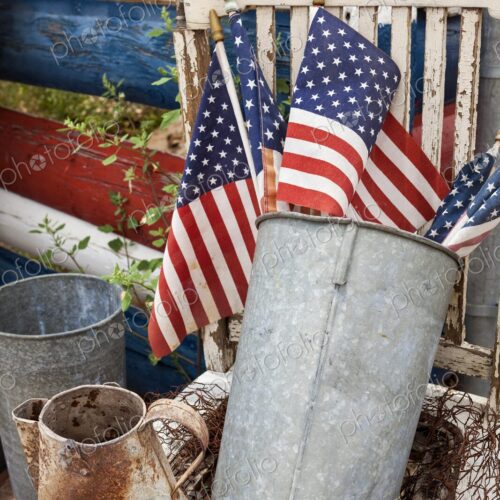 American flags in a weathered metal bucket sit on a rustic chair beside aged farm…