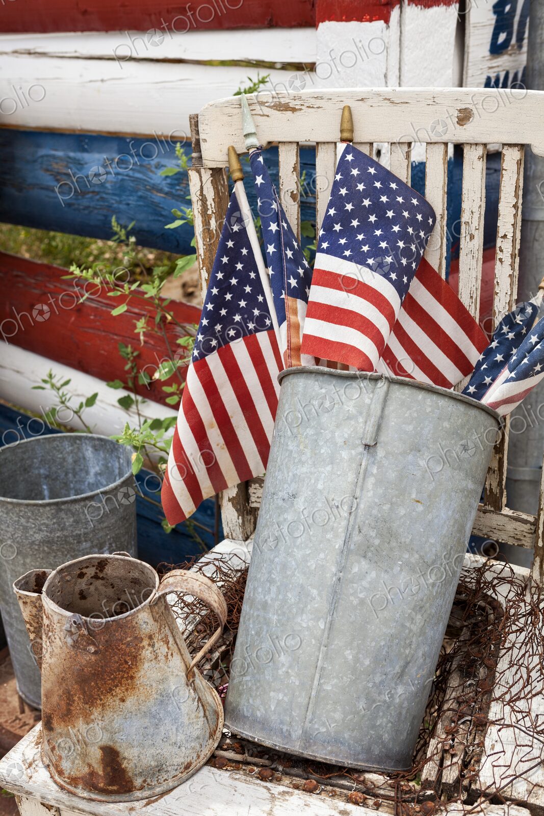 American flags in a weathered metal bucket sit on a rustic chair beside aged farm…