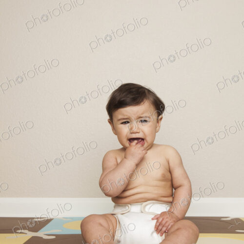 An 8 month old baby boy sitting on the floor in a diaper crying. An 8 month old baby boy sitting on the floor in a diaper crying.