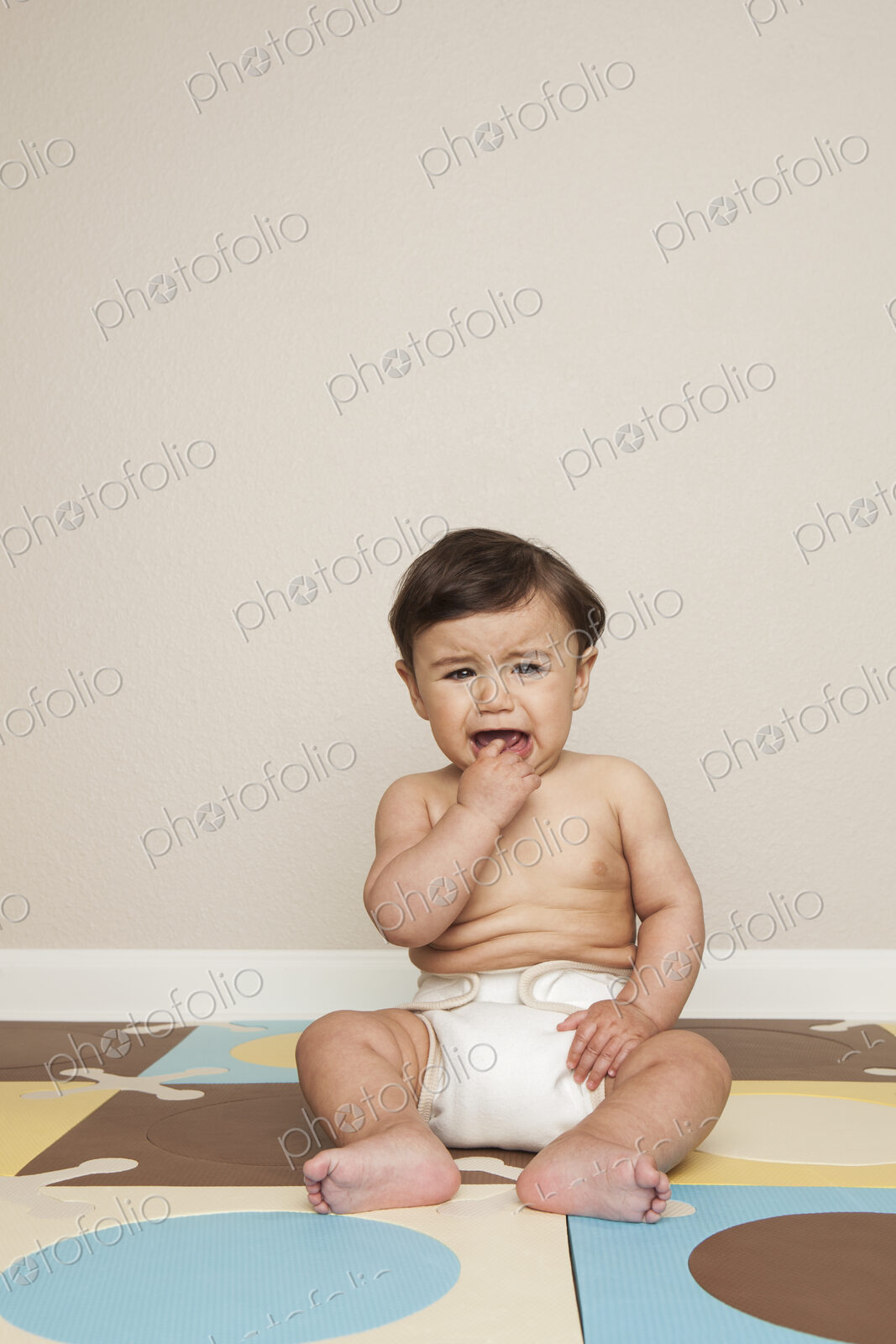 An 8 month old baby boy sitting on the floor in a diaper crying.