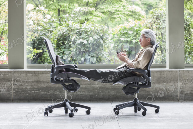 An Asian businessman checking his phone, sitting in a chair and resting his feet in…
