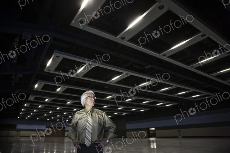 An Asian businessman standing in a dimly lit and dark exhibition area in a convention…