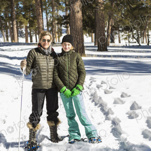 An adult woman and teenage girl in snow shoes in woodland holding ski poles