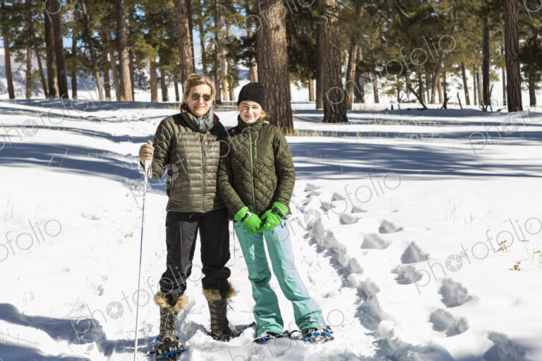 An adult woman and teenage girl in snow shoes in woodland holding ski poles