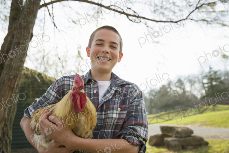 An animal sanctuary. A young boy holding a chicken with brown feathers and a red coxcomb.