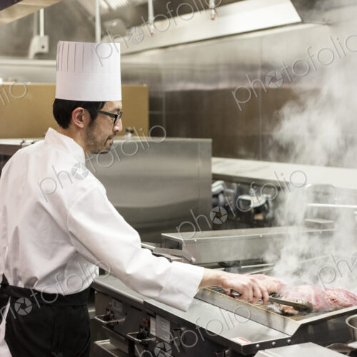 An asian chef working with fresh meat on the grill in a commercial kitchen,