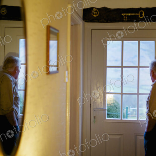 An elderly man standing by a door looking out through the window panes. An elderly man standing by a door looking out through the window panes.