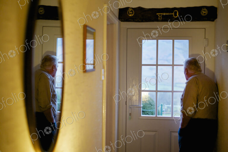 An elderly man standing by a door looking out through the window panes.
