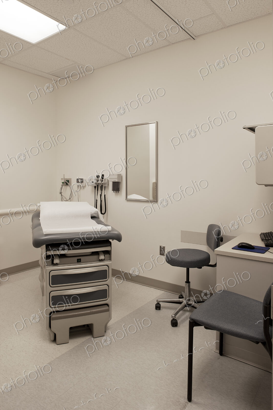 An examination room in a hospital, with a treatment table, patient bed, a stool and chair.