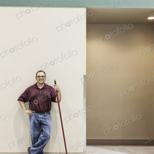 An hispanic workman standing in a large interior space with a broom.