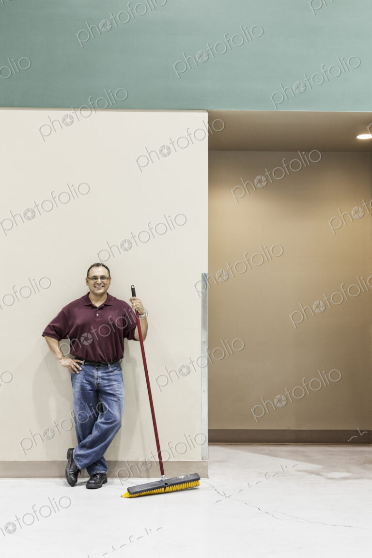 An hispanic workman standing in a large interior space with a broom.