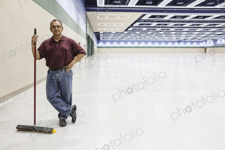 An hispanic workman standing in a large interior space with a broom.