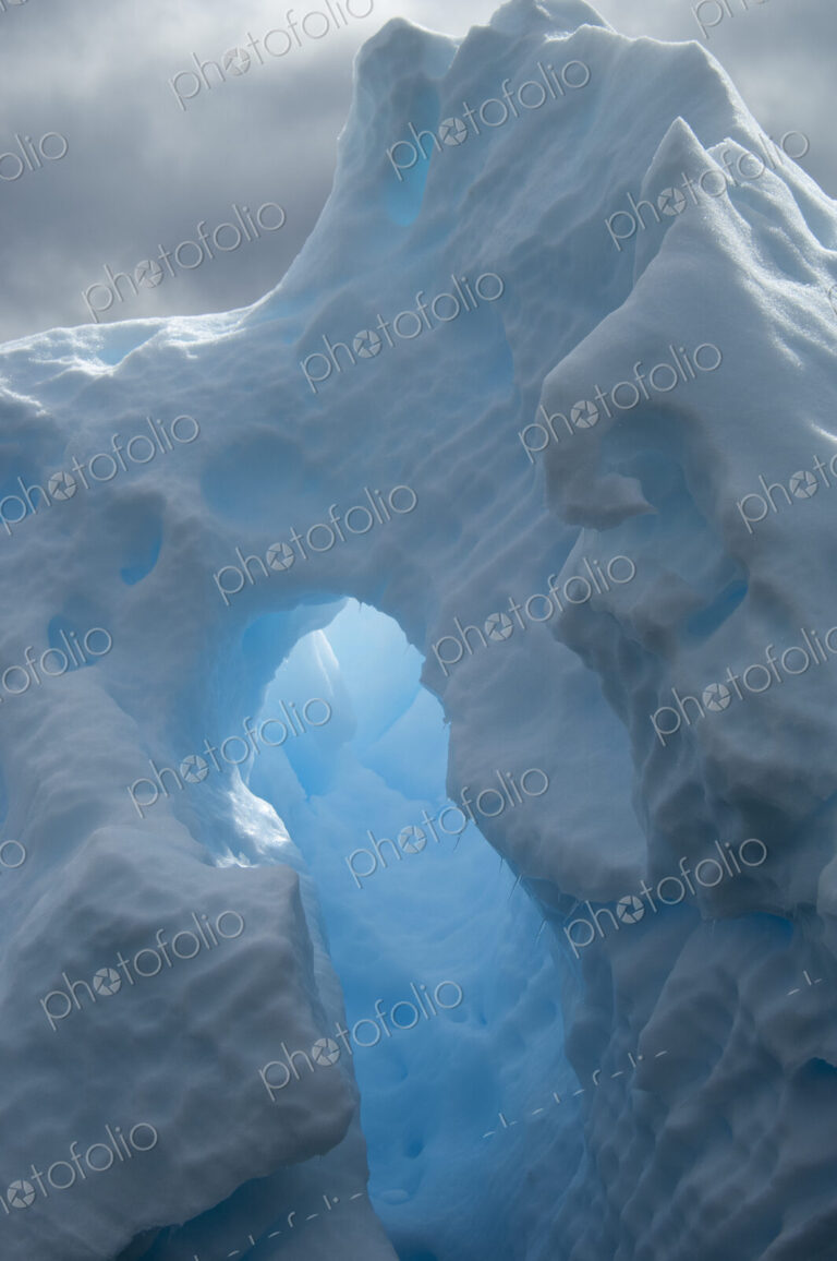 An iceberg with a natural arch and translucent glow, with sunlight shining through the…