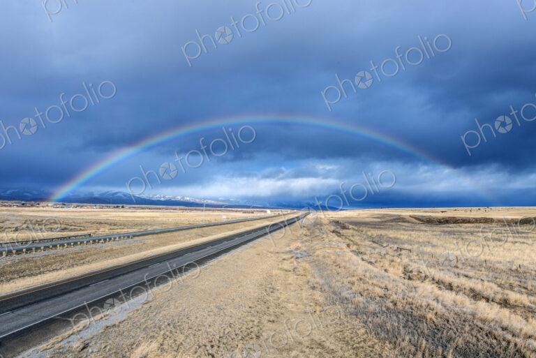 An interstate road reaching to the horizon through a flat landscape, rainbow above the…