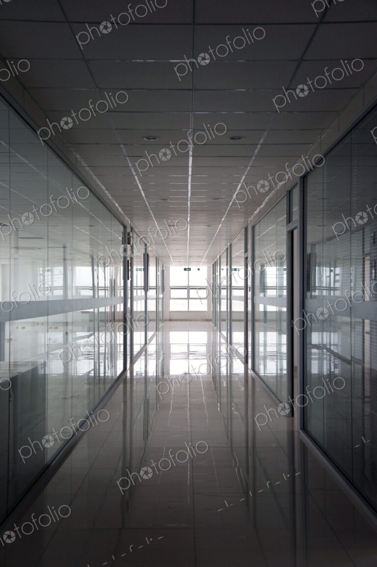 An office interior, straight wide corridor with reflective surfaces on walls and floor…