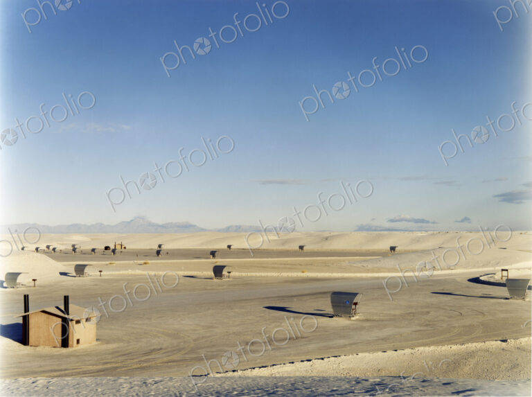 An open space a desert landscape, with a small cabin and sun shelters, surrounded by…