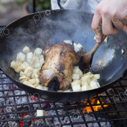 An outdoor cookout, a game bird in a pan surrounded by mirepoix of vegetables above a… An outdoor cookout, a game bird in a pan surrounded by mirepoix of vegetables above a…