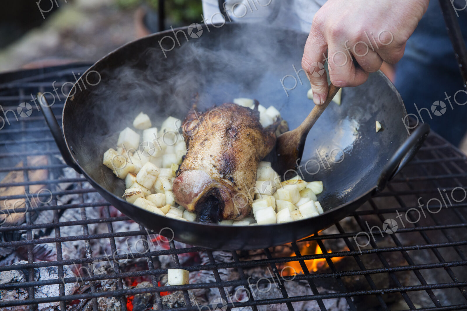 An outdoor cookout, a game bird in a pan surrounded by mirepoix of vegetables above a…