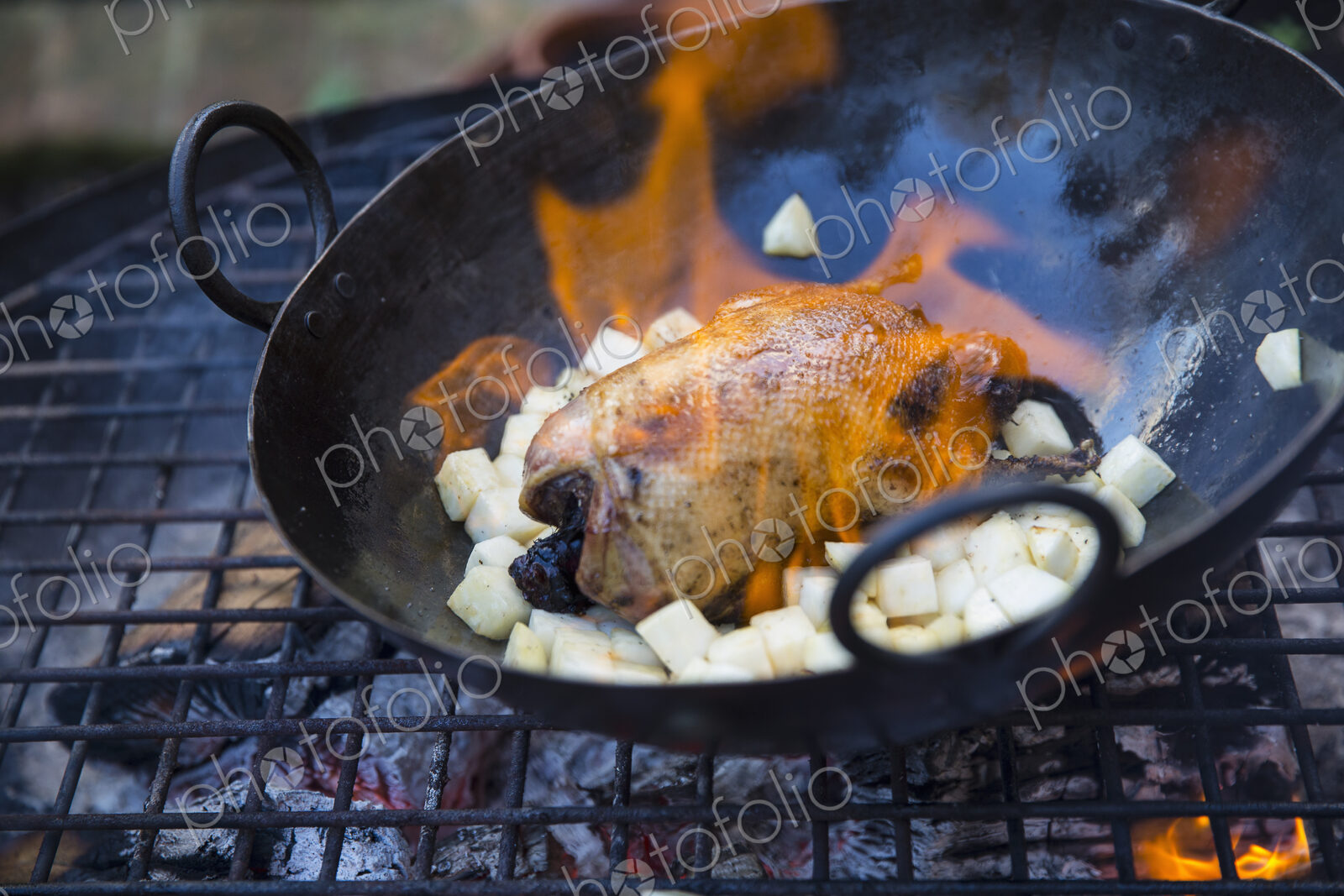 An outdoor cookout, a game bird in a pan with flame licking around the carcass,…