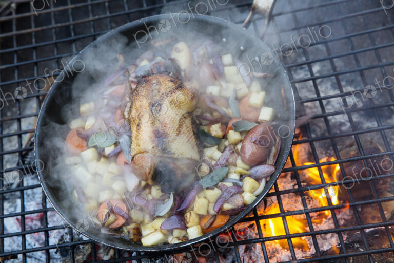 An outdoor cookout, game birds in a pan above a glowing fire.