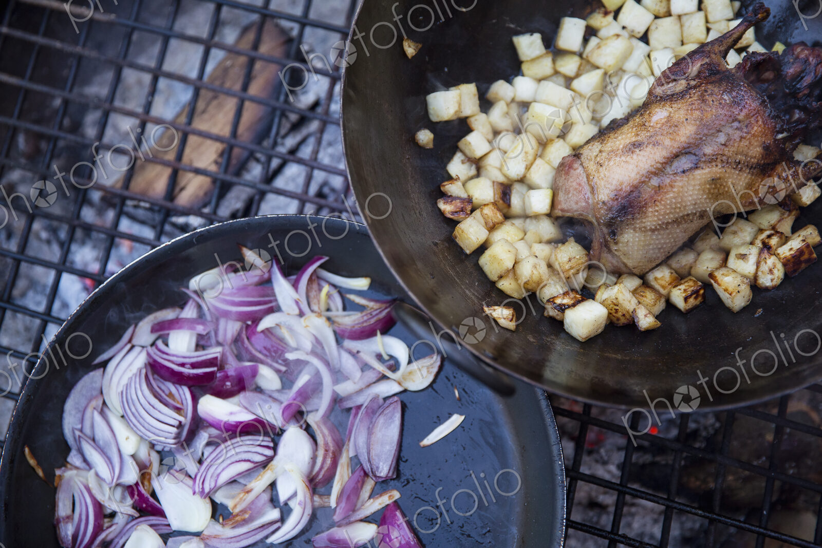 An outdoor cookout, two frying pans with vegetables and a small game bird.