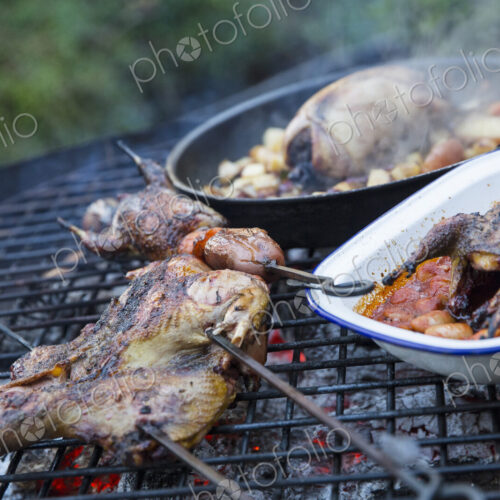 An outdoor cookout, two small game birds in a pan and one being barbequeued above hot… An outdoor cookout, two small game birds in a pan and one being barbequeued above hot…