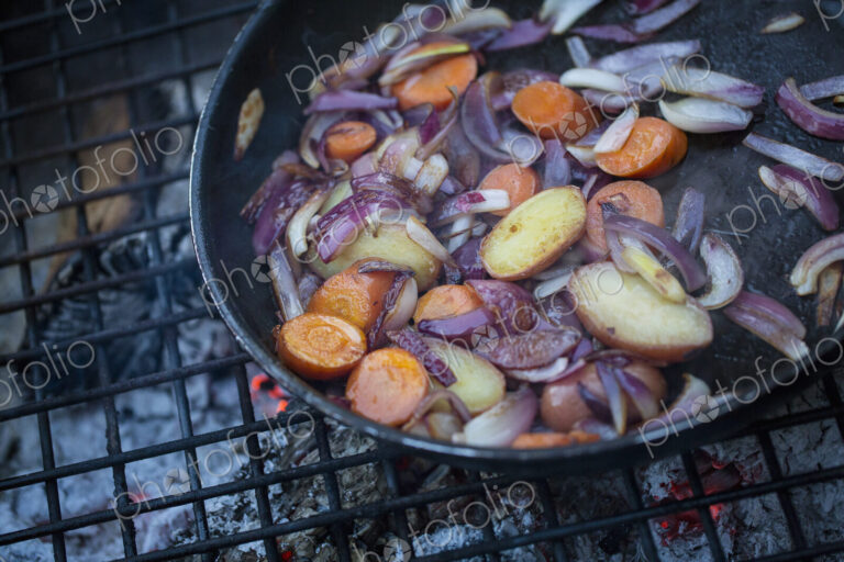 An outdoor cookout, vegetables in a frying pan above a glowing fire.