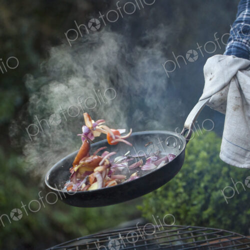 An outdoor cookout. A man holding a frying pan sauteeing vegetables above an open fire. An outdoor cookout. A man holding a frying pan sauteeing vegetables above an open fire.