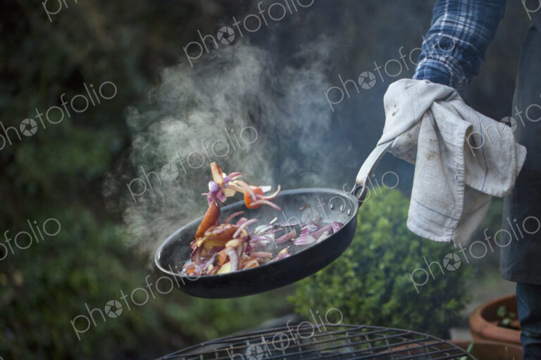 An outdoor cookout. A man holding a frying pan sauteeing vegetables above an open fire.