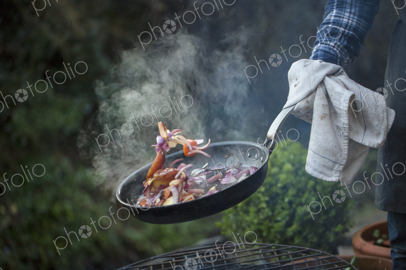 An outdoor cookout. A man holding a frying pan sauteeing vegetables above an open fire.