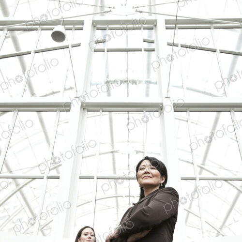 Asian business woman and caucasian business woman in suit coats standing in a glass… Asian business woman and caucasian business woman in suit coats standing in a glass…