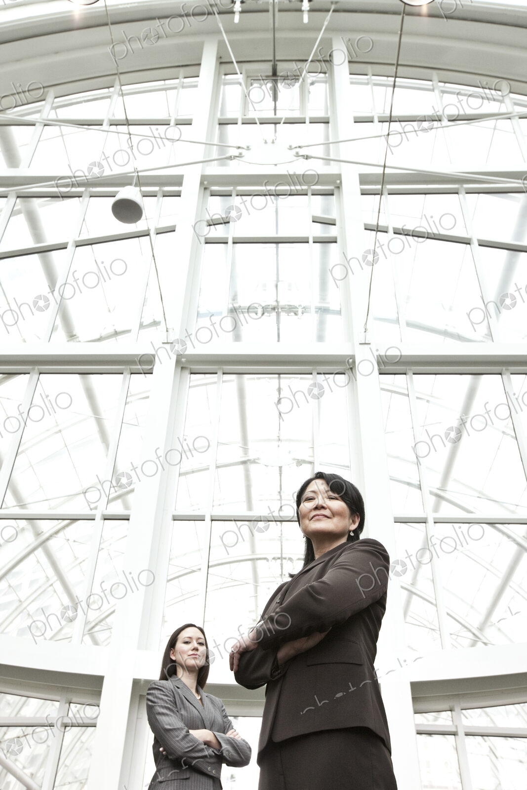 Asian business woman and caucasian business woman in suit coats standing in a glass…
