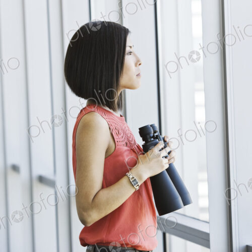 Asian business woman at a business lobby window holding binoculars.