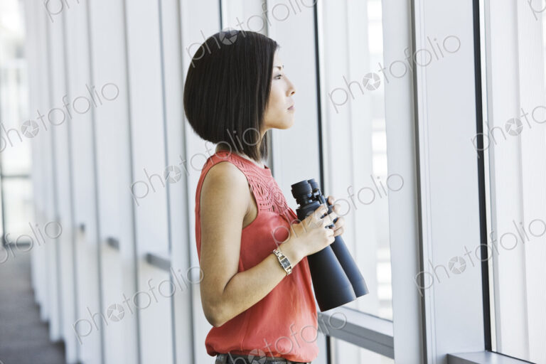 Asian business woman at a business lobby window holding binoculars.