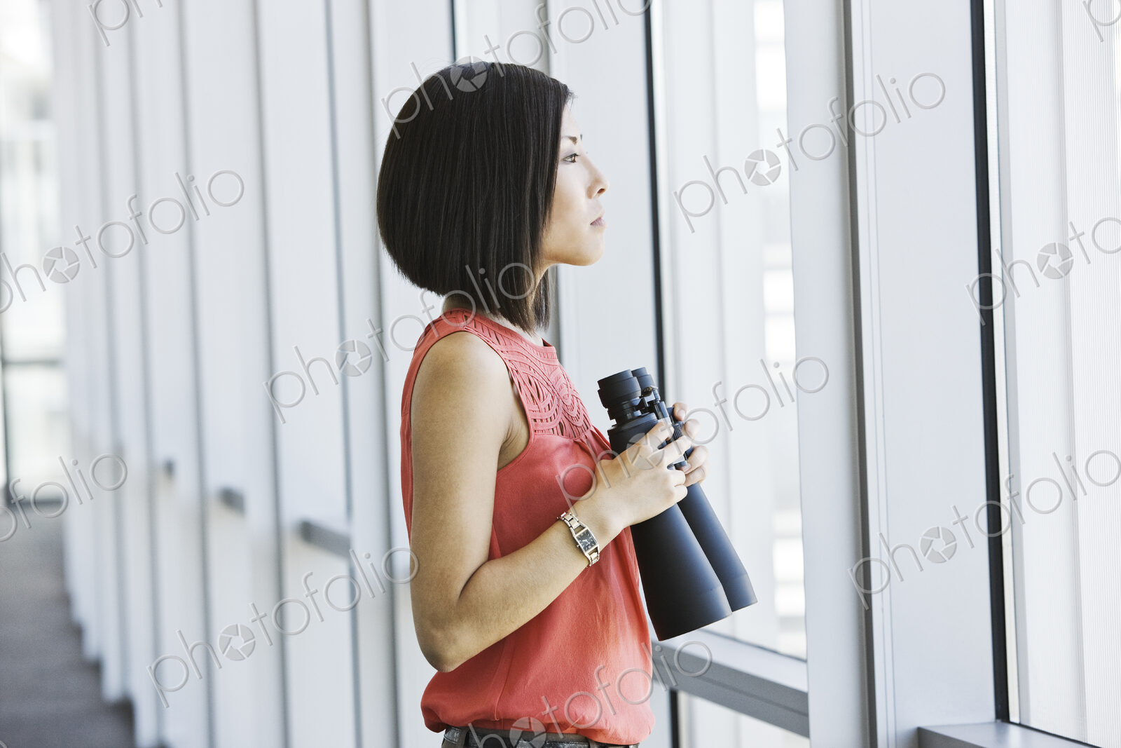 Asian business woman at a business lobby window holding binoculars.