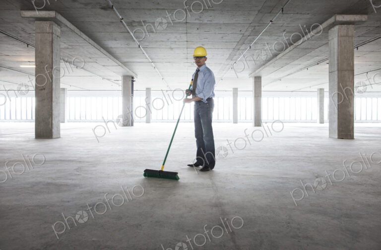 Asian businessman cleaning up with a broom in a large empty raw office space.