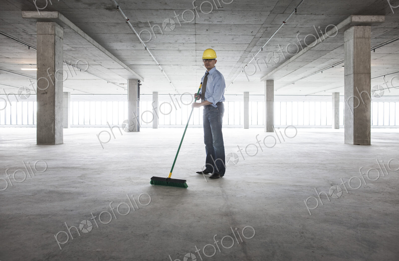 Asian businessman cleaning up with a broom in a large empty raw office space.