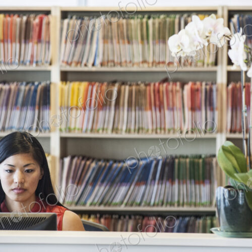 Asian female working in a medical office with file folders in the background Asian female working in a medical office with file folders in the background