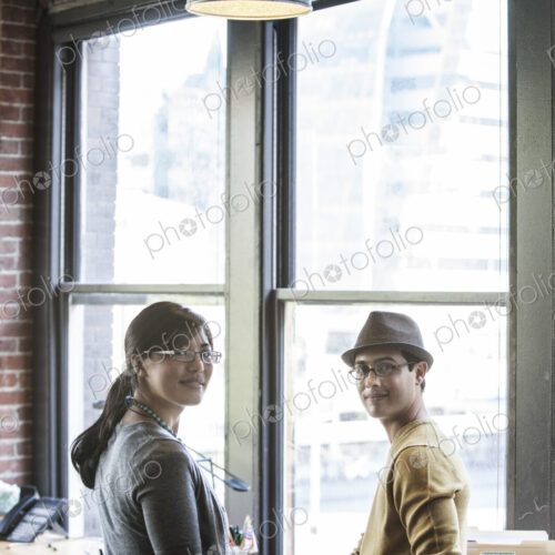 Asian woman and Hispanic man together next to large windows in a creative office. Asian woman and Hispanic man together next to large windows in a creative office.