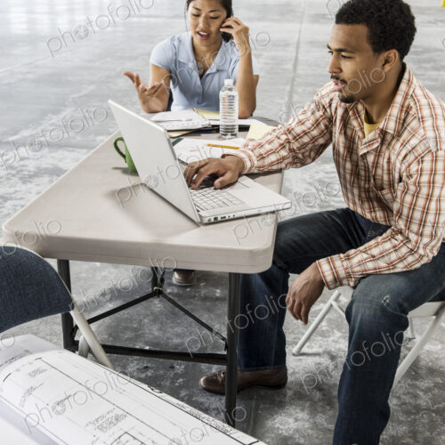 Asian woman and black man sitting at a desk in front of loading dock doors working on… Asian woman and black man sitting at a desk in front of loading dock doors working on…