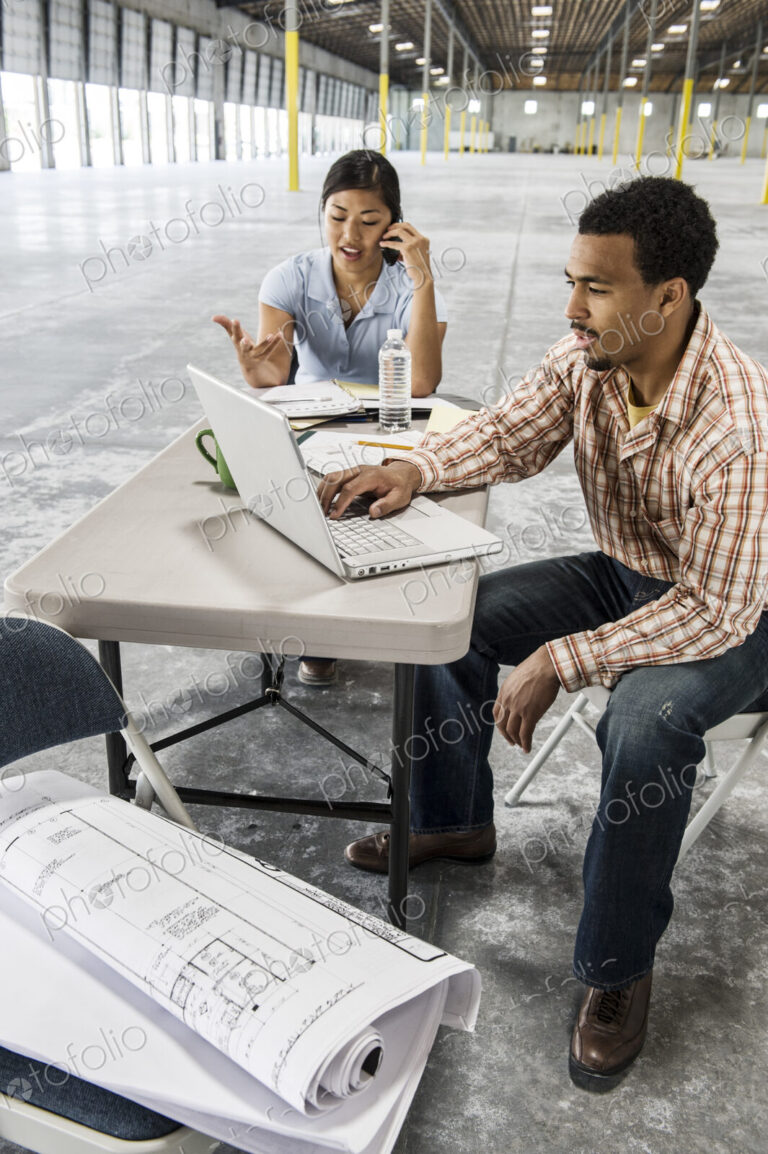 Asian woman and black man sitting at a desk in front of loading dock doors working on…