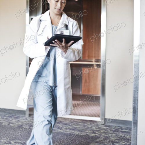 Asian woman doctor walking in a hospital hallway while working on a notebook computer.