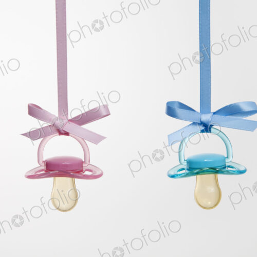 Baby pacifiers hanging from ribbons against a plain background.
