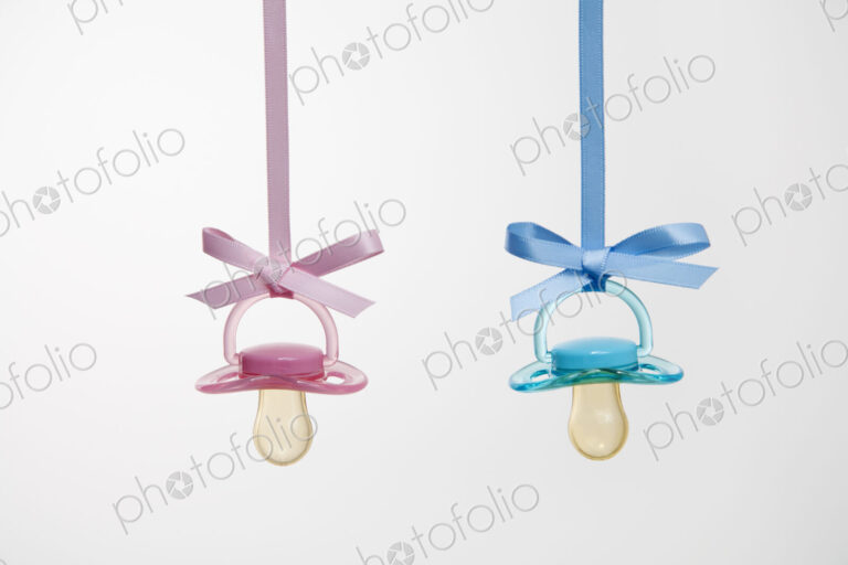 Baby pacifiers hanging from ribbons against a plain background.