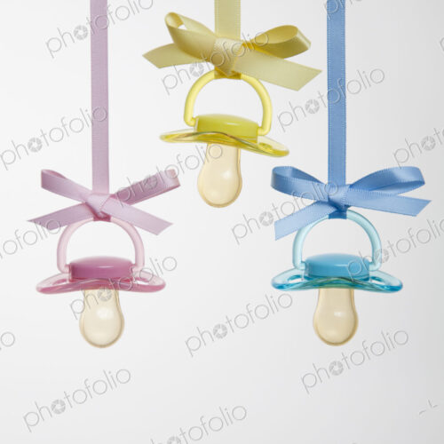 Baby pacifiers hanging from ribbons against a plain background.