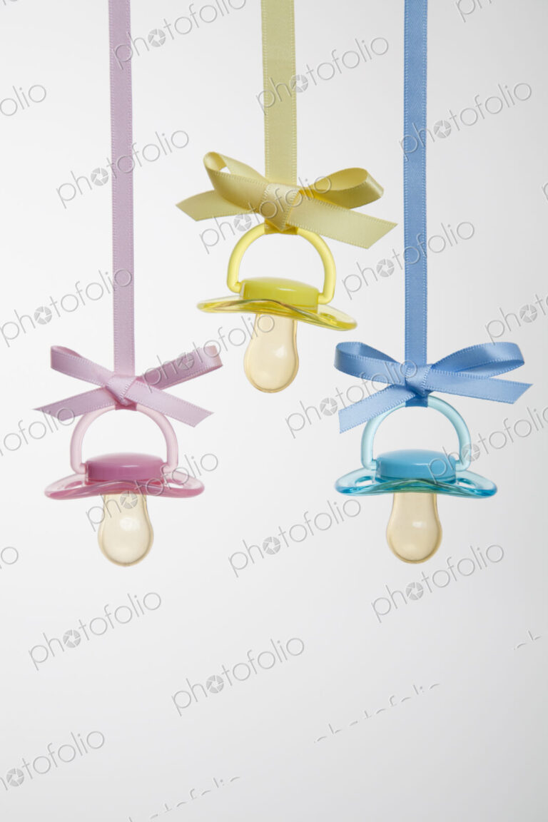 Baby pacifiers hanging from ribbons against a plain background.