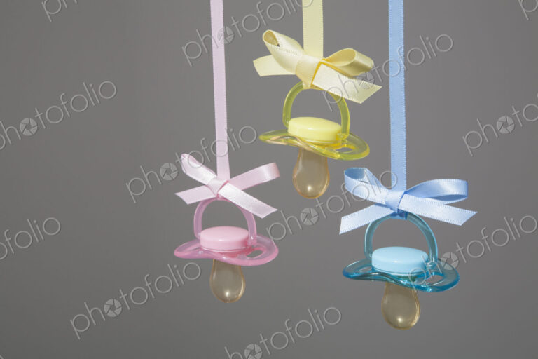 Baby pacifiers hanging from ribbons against a plain background.