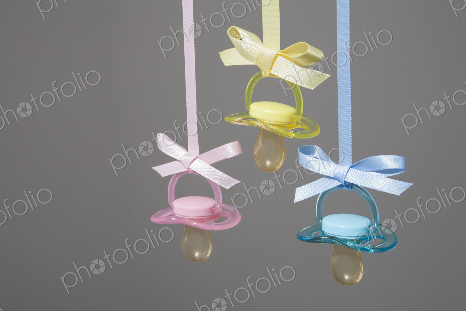 Baby pacifiers hanging from ribbons against a plain background.