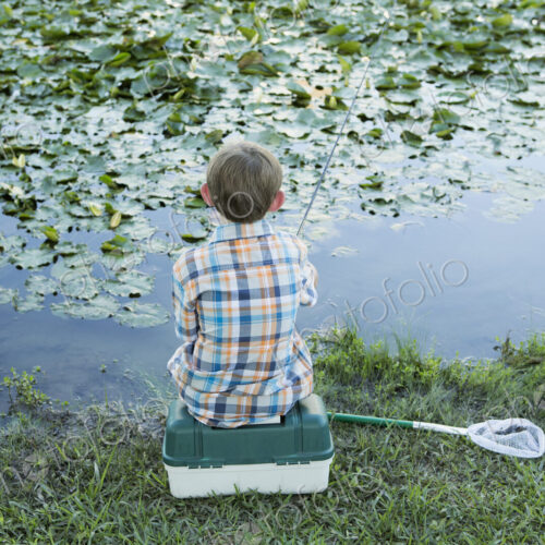 Back view of a young boy sitting on his baitbox, fishing in the river.