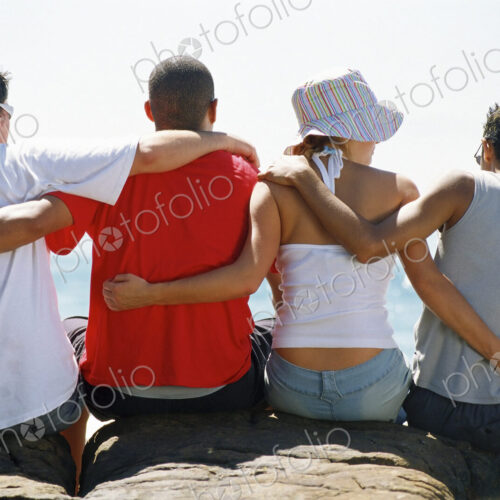 Back view of four friends, with arms around each other, sitting in a row. Back view of four friends, with arms around each other, sitting in a row.